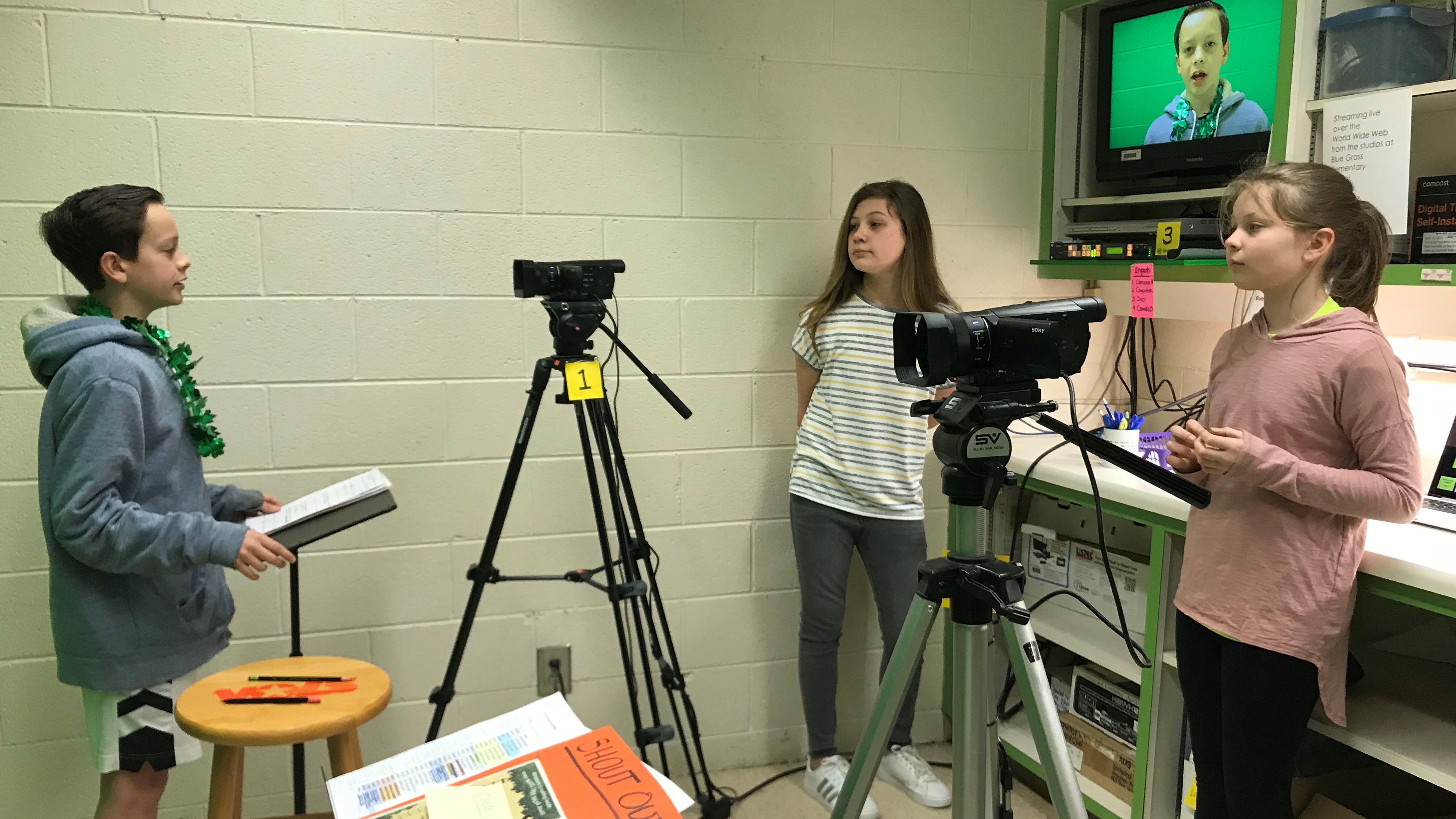 Micah Kohring (left) anchors the morning newscast at Blue Grass Elementary School, as Anna Sergent (center) and Rachel Hendon monitor the cameras.. 